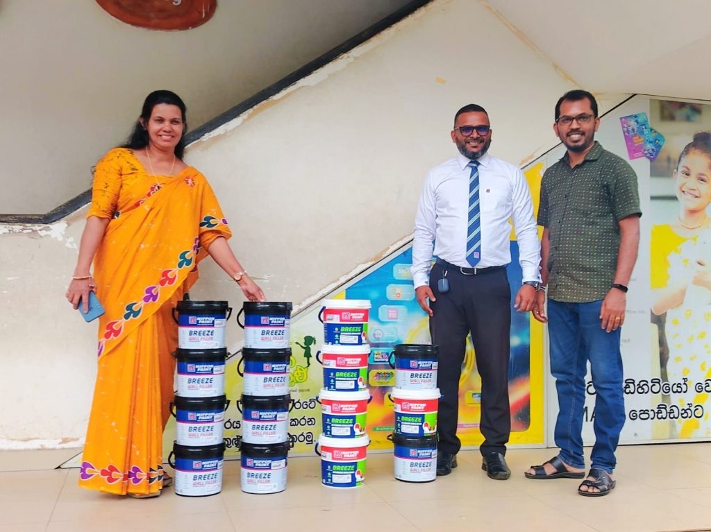 Three people standing next to stacks of Nippon Paint buckets in a school hallway.