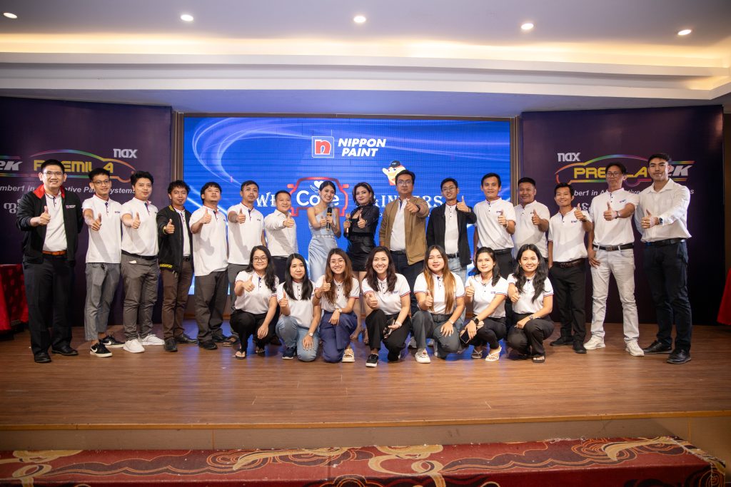 A large group of attendees and team members posing enthusiastically on stage at the We Care Painter Automotive Refinishing event.