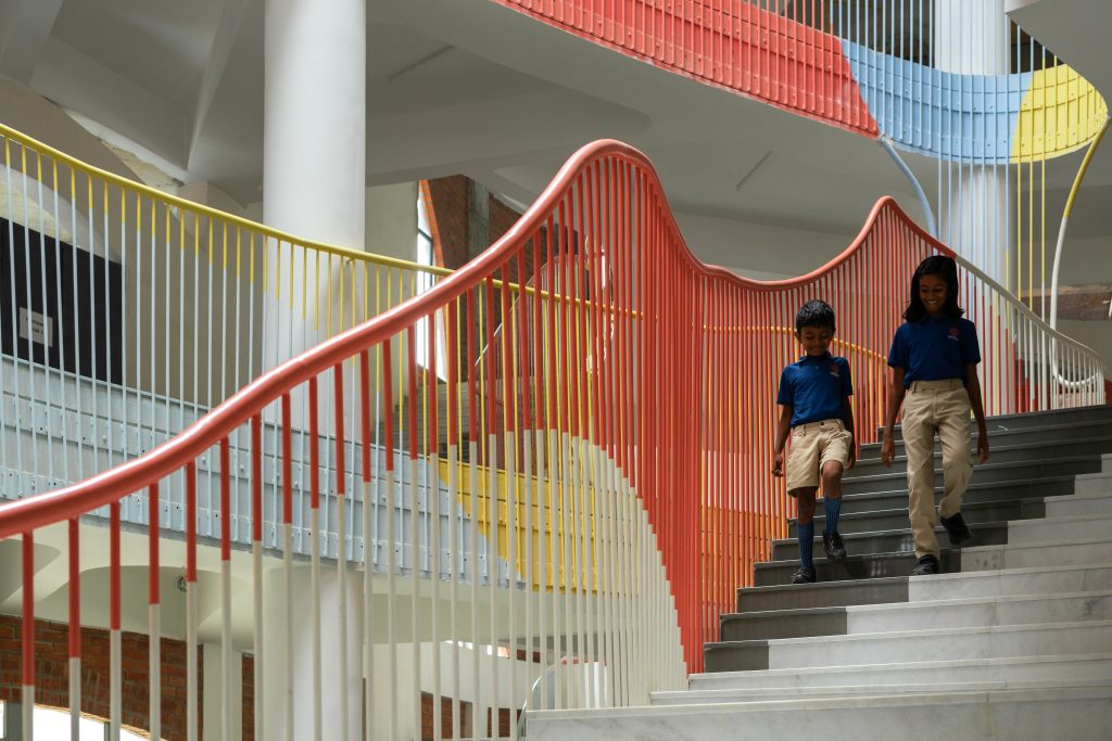 Two young students walking down a curved staircase enclosed by bright red railings.