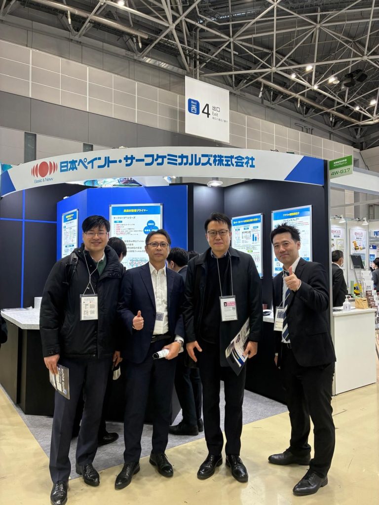 Four NCK team members standing in front of their blue exhibition booth at Tokyo Big Sight.