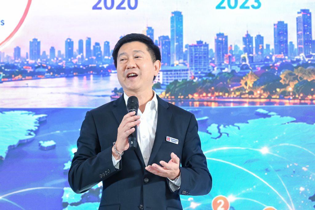 General Manager Watchara Siriritthichai smiling and delivering an engaging speech on stage against a vibrant cityscape backdrop.