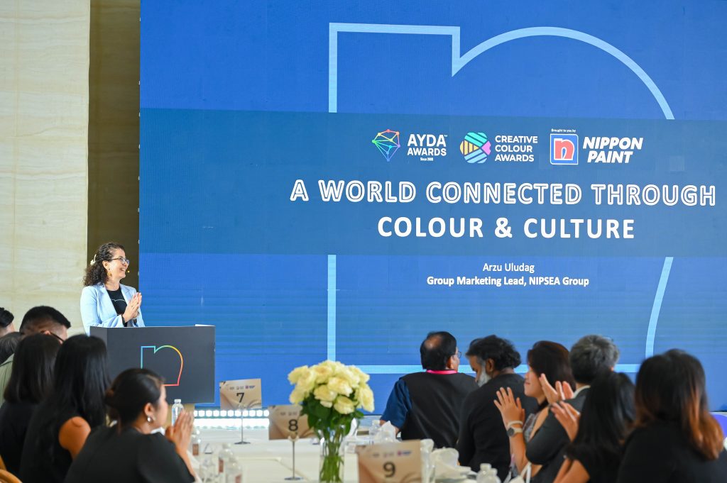 Group Marketing Lead Arzu Uludag speaking on stage with a digital presentation slide reading "A World Connected Through Colour & Culture".