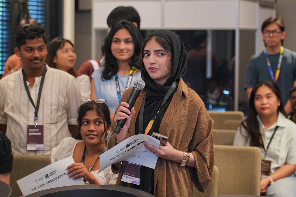 A young woman in a hijab speaking into a microphone while holding a bingo paper.