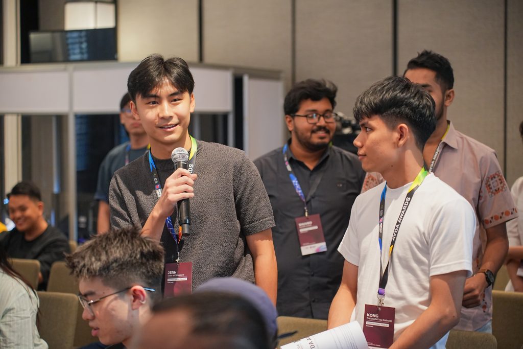 A young male finalist standing and confidently speaking into a microphone among the audience.