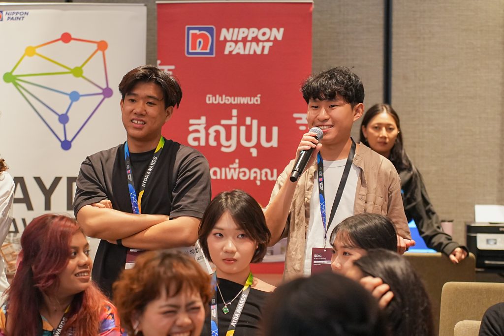 A smiling young man standing to speak into a microphone at the AYDA event.