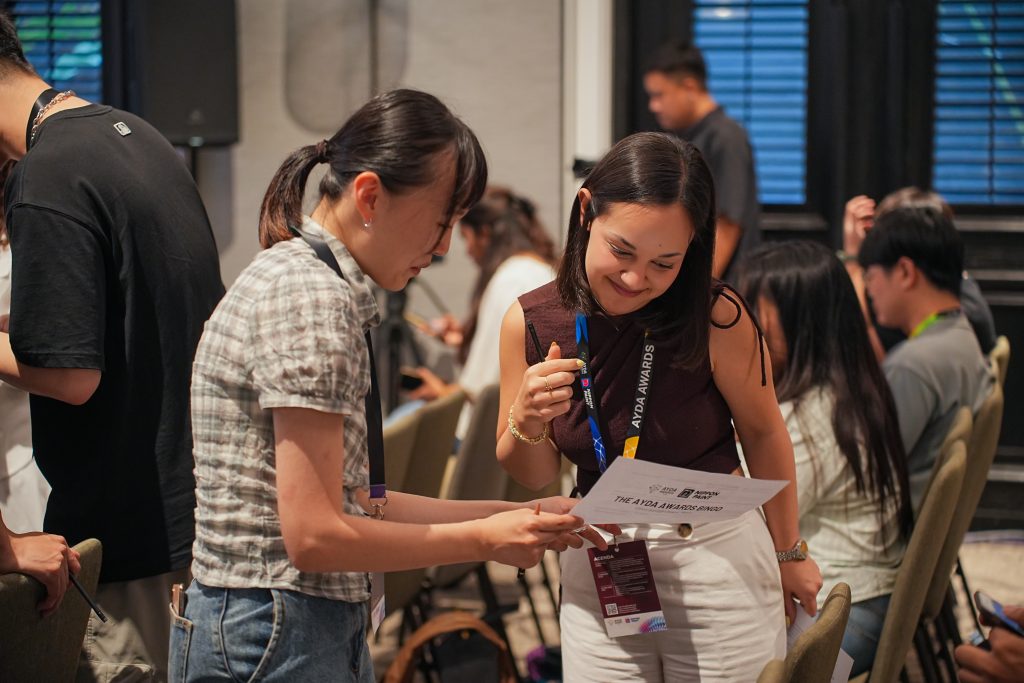 Two female participants smiling and collaborating over an AYDA Awards Bingo sheet.