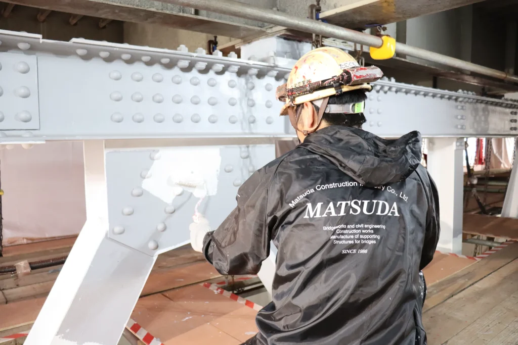 A construction worker in a black Matsuda jacket painting a large steel bridge beam with white paint using a roller.
