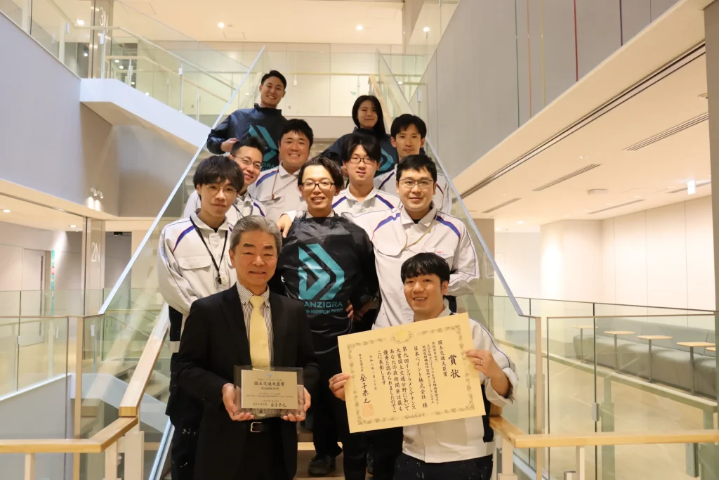 A group of smiling Japanese workers and officials posing on a bright staircase, holding a commemorative plaque and a certificate.