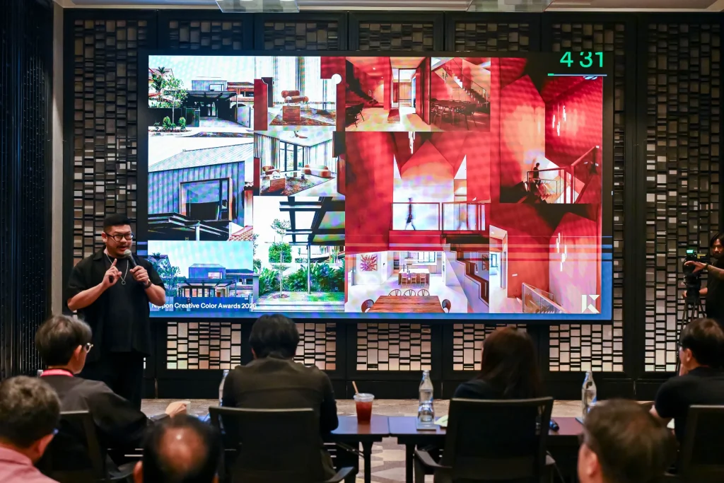 A male designer in a black shirt holding a microphone, presenting his architectural project on a large digital screen to a seated audience.