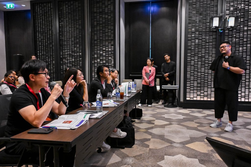 A panel of judges sitting at a table, actively interacting with a standing male presenter.