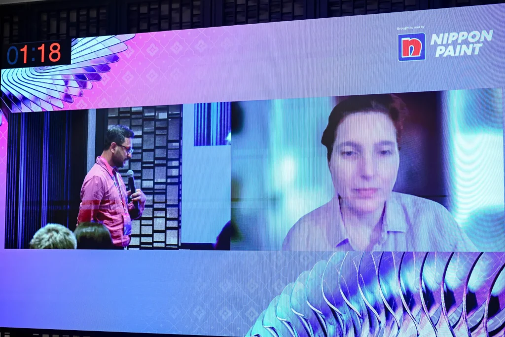 A large projection screen at an event displaying a man speaking on stage alongside a video call feed of Francesca Heathcote Sapey, Partner and Managing Director at Teresa Sapey + Partners.