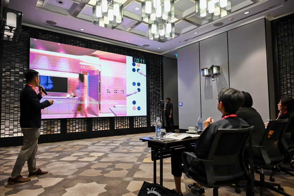 A man standing on stage, presenting a vibrant interior design project to the seated judges.
