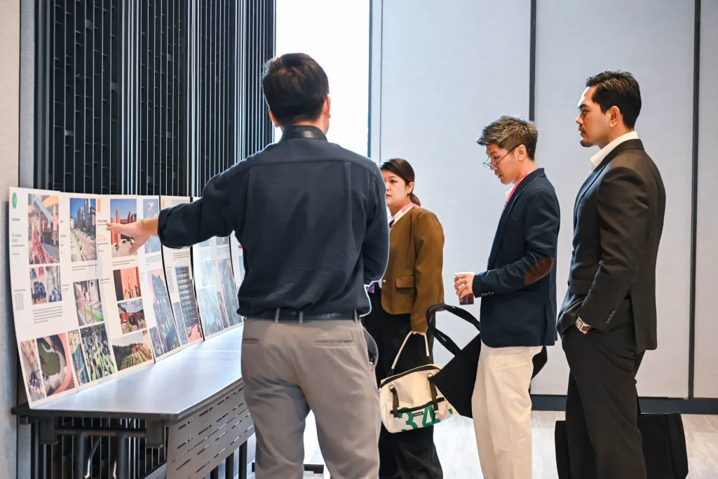 A group of attendees standing around a table, actively pointing at and discussing printed architectural presentation boards.