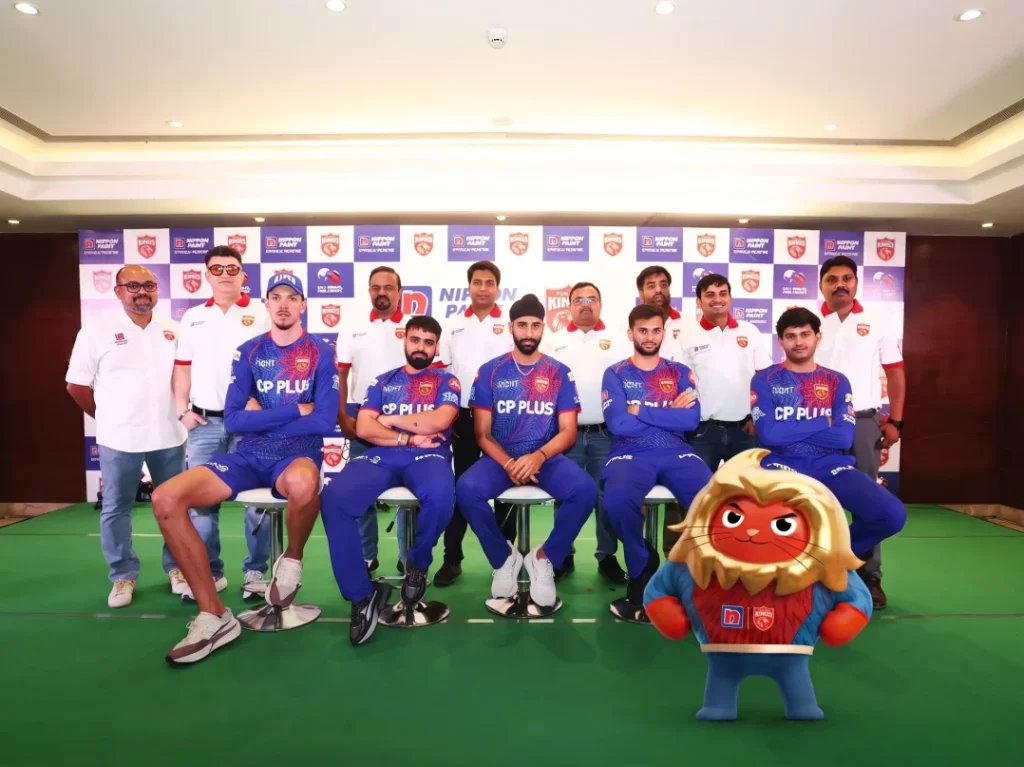 Punjab Kings cricket players sitting with staff and a mascot at a press event.