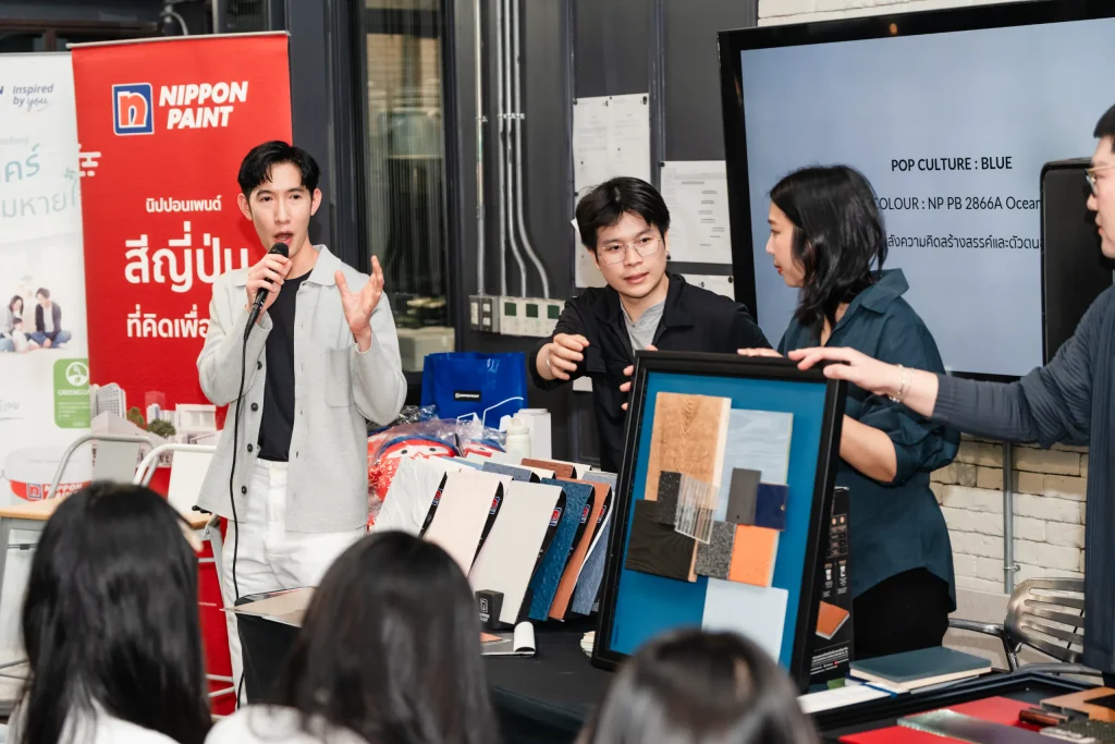A speaker talks into a microphone while a colleague presents a board of textured materials.