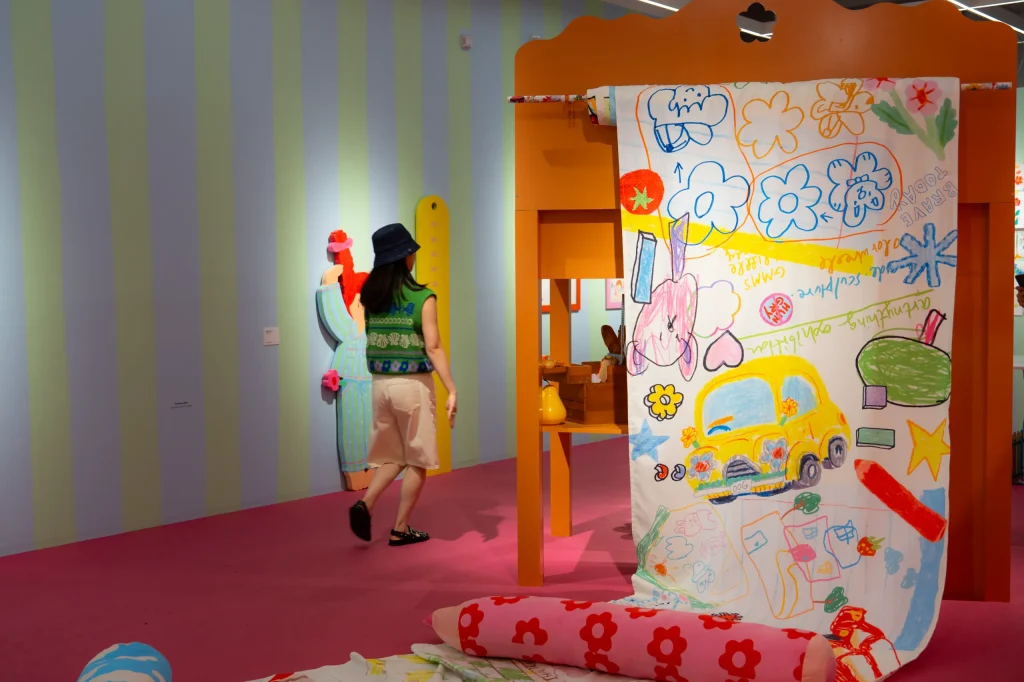 A person walking past a large, hanging scroll covered in colourful, childlike crayon drawings.
