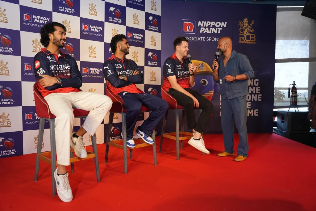 Three RCB cricket players sit on stools answering questions from a host on stage.