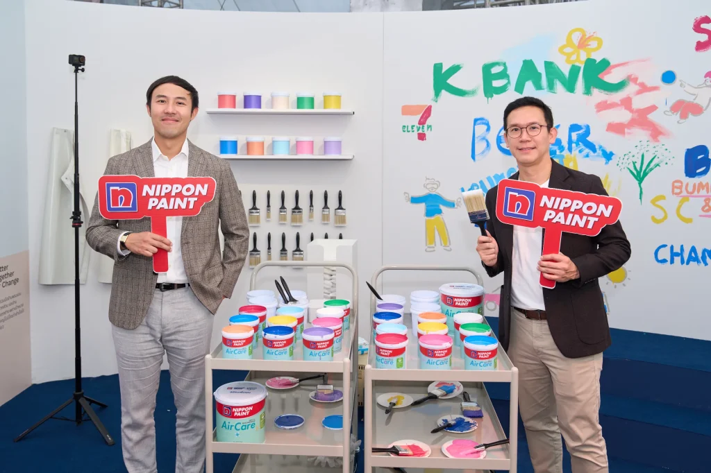 Two men in smart casual attire hold Nippon Paint signs next to paint supplies.